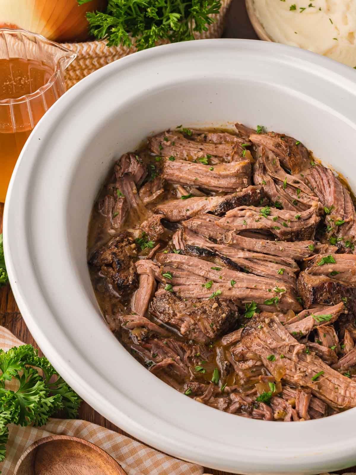 Juicy shredded beef in a slow cooker surrounded by cooking juices and topped with parsley.