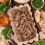 Shredded beef on a platter served with mashed potatoes, peas, bread, and fresh herbs for dinner.