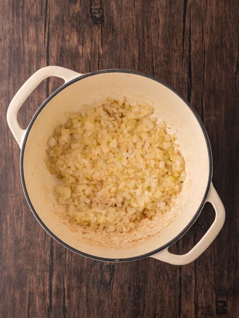 Diced onions sautéing in a cream-colored Dutch oven on a wooden surface for pumpkin coconut curry soup.