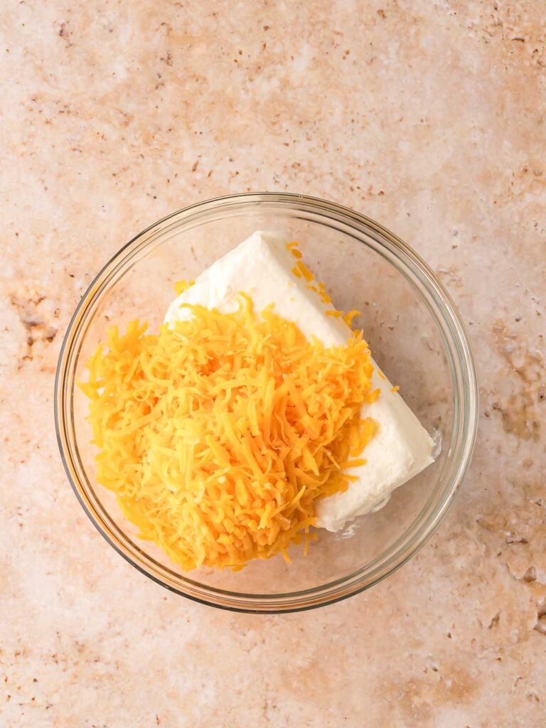 Glass mixing bowl with softened cream cheese and shredded cheddar cheese, ready to be mixed for pumpkin-shaped cheese balls.