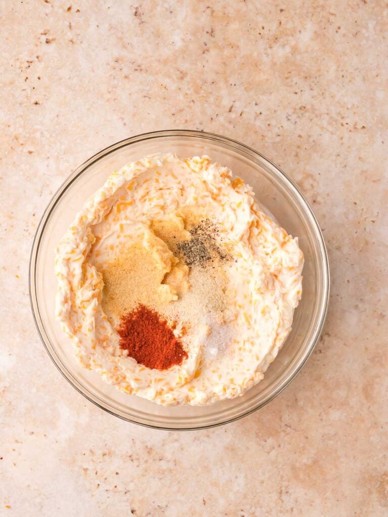 Seasoned cheese mixture in a glass bowl with garlic powder, onion powder, smoked paprika, salt, and pepper added on top before stirring.