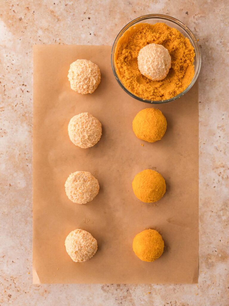 Cheese balls in the process of being coated with crushed cheese crackers, with some already covered and others still plain.