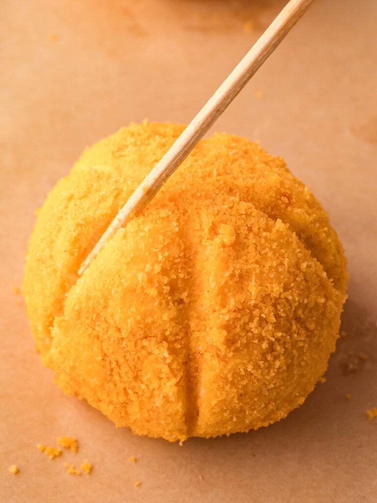 Close-up of a cheese ball being shaped with vertical pumpkin-like lines using a skewer before adding toppings.