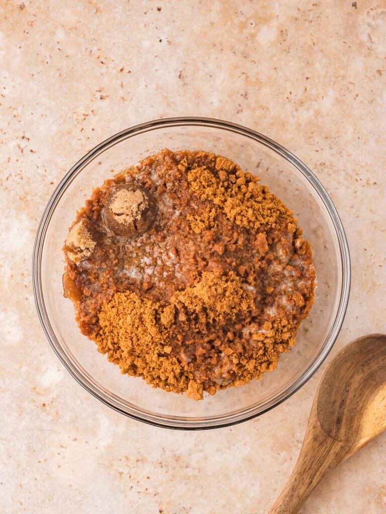 Mixing crushed Biscoff cookies, brown sugar, and melted butter in a glass bowl for no-bake pumpkin cheesecake bars crust.