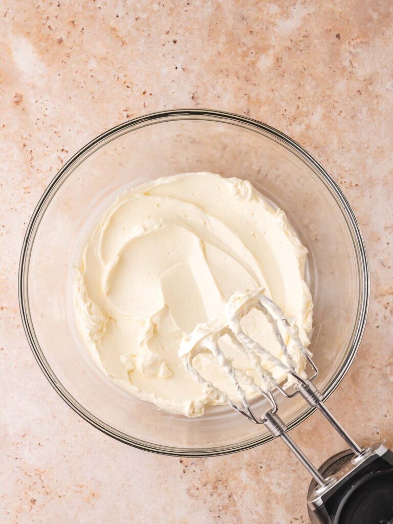 Whipped cream cheese in a glass mixing bowl with beaters, step for making no-bake pumpkin cheesecake bars filling.
