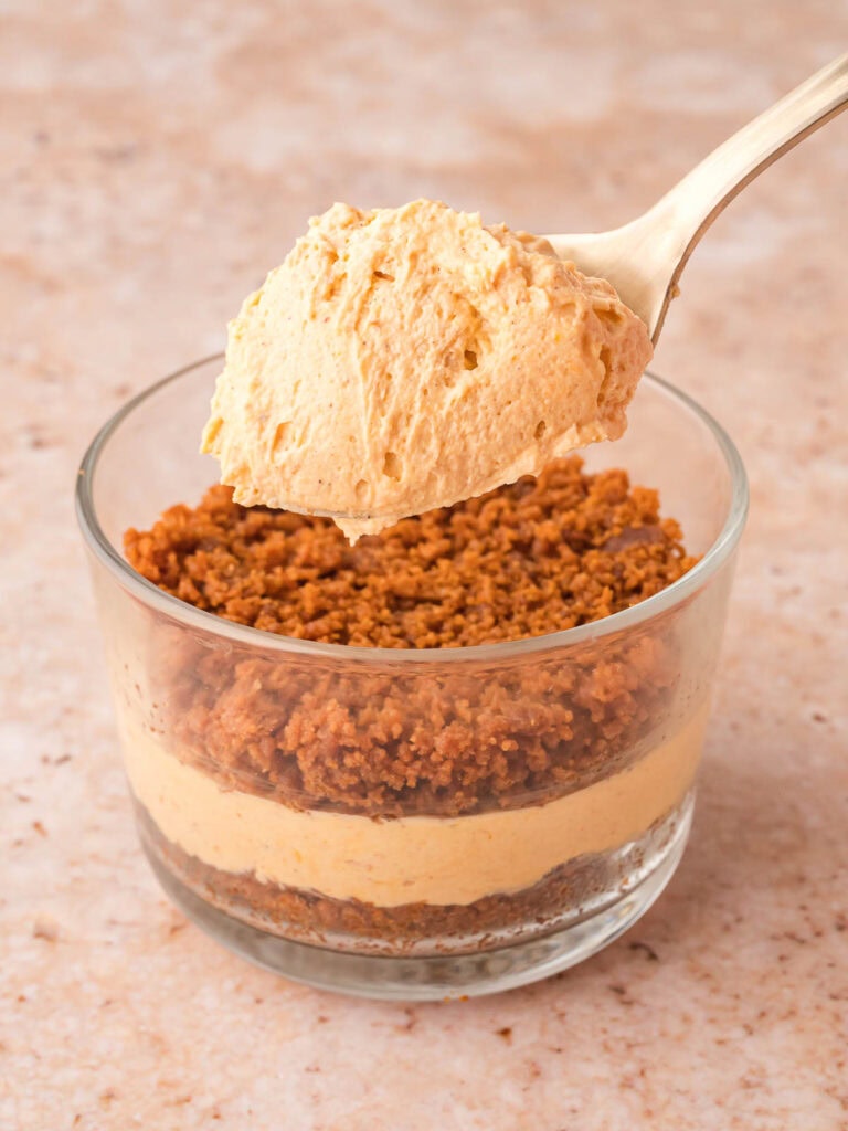 Spoon adding pumpkin cheesecake filling on top of the crust layer in a glass for pumpkin pie cheesecake cups.