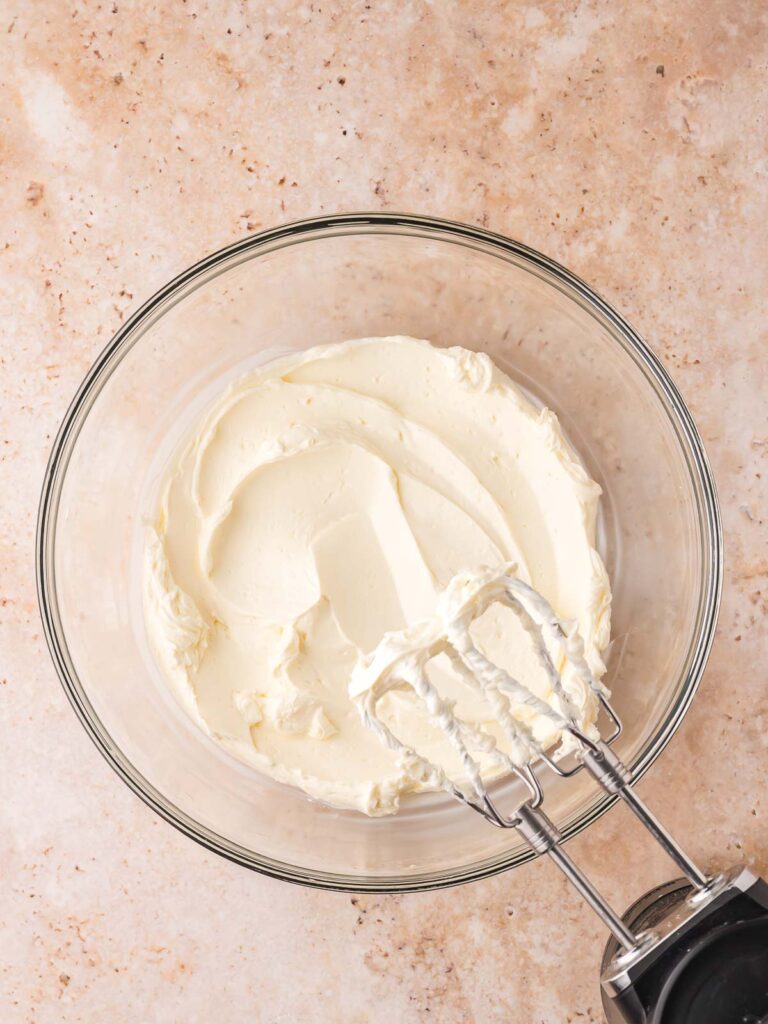 Whipped cream cheese in a glass mixing bowl with beaters, step for making pumpkin pie cheesecake cups filling.