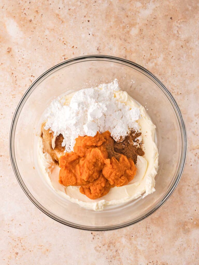 Cream cheese mixture with pumpkin purée, powdered sugar, and spices added to the bowl for pumpkin pie cheesecake cups.