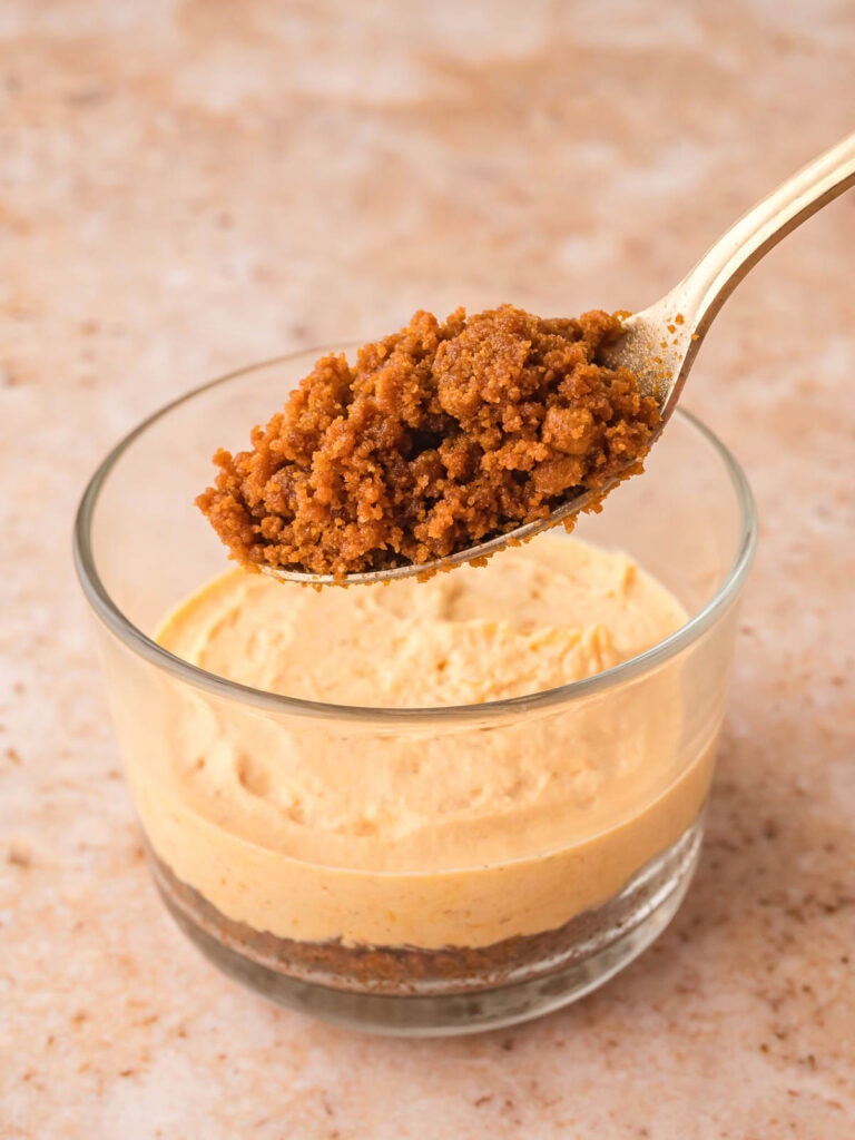 Spoon adding another layer of Biscoff cookie crust over the pumpkin cheesecake filling in a glass cup.