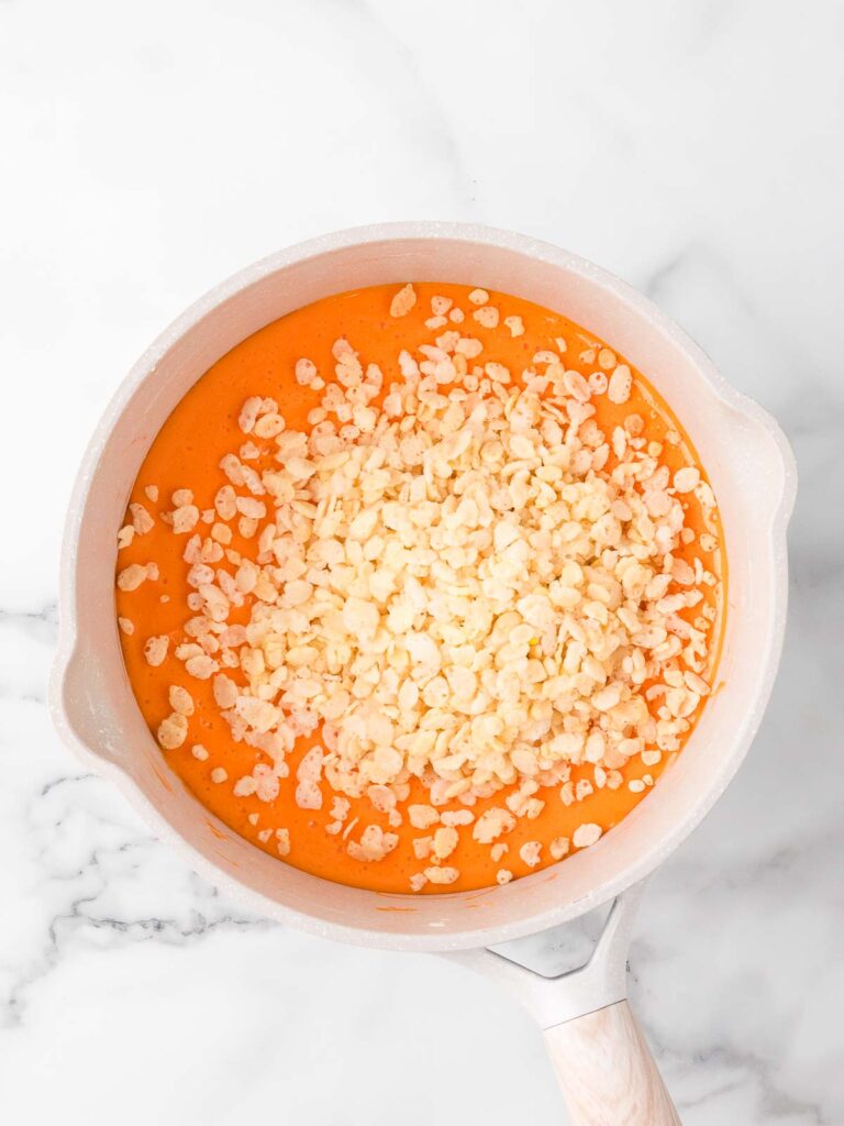 Mixing Rice Krispies cereal into melted orange marshmallow mixture in a saucepan.