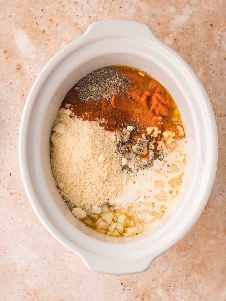 Uncooked ingredients in a slow cooker including pumpkin purée, Parmesan, garlic, heavy cream, and spices before mixing.