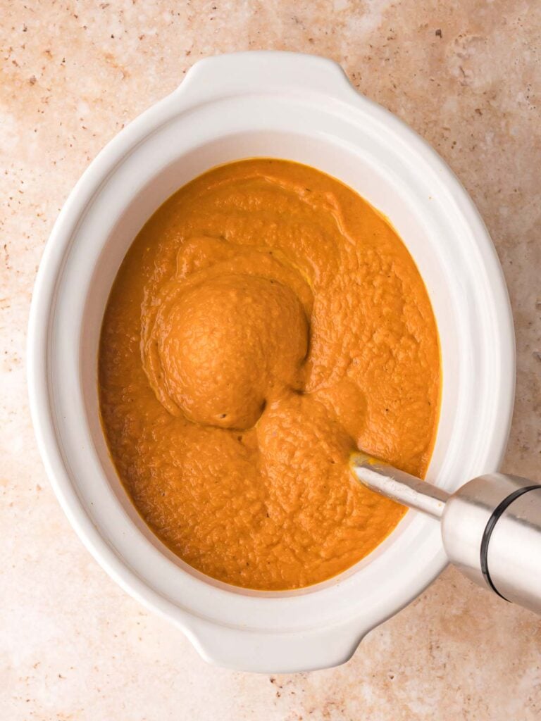 Pumpkin soup being blended directly in the slow cooker with an immersion blender until smooth.