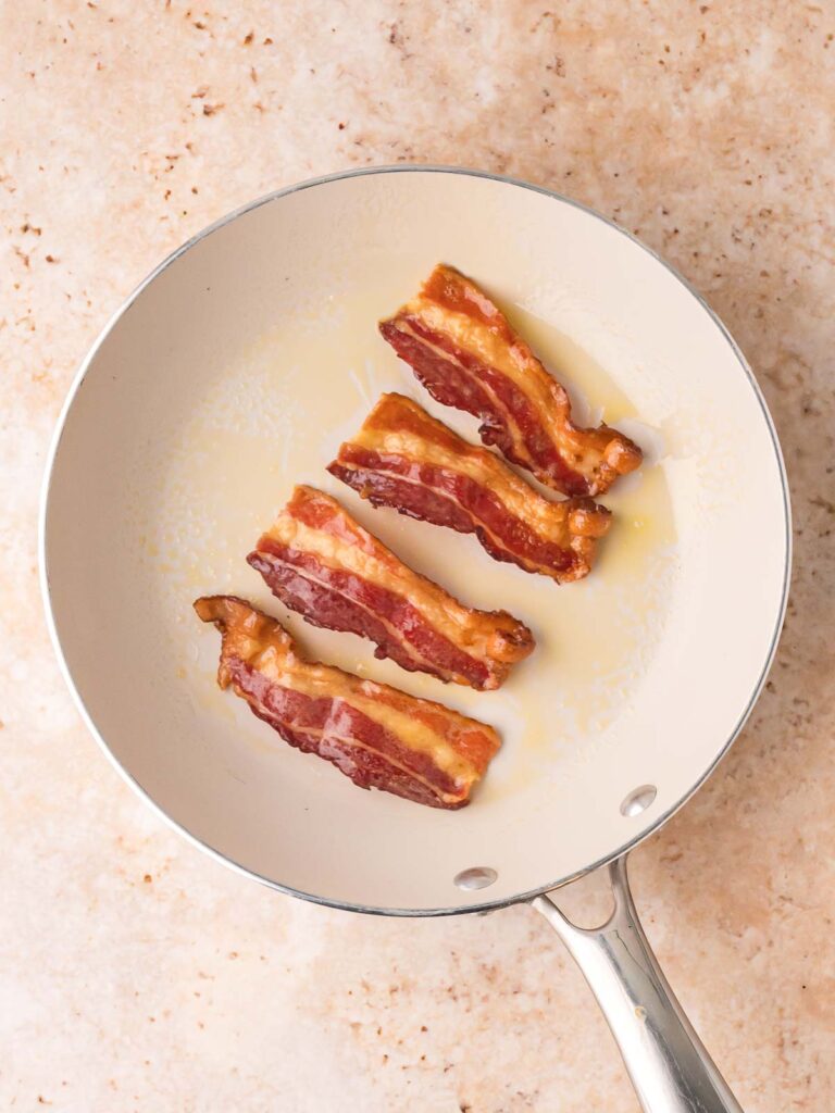 Four strips of thick-cut bacon sizzling in a nonstick skillet, partially cooked for soup topping.