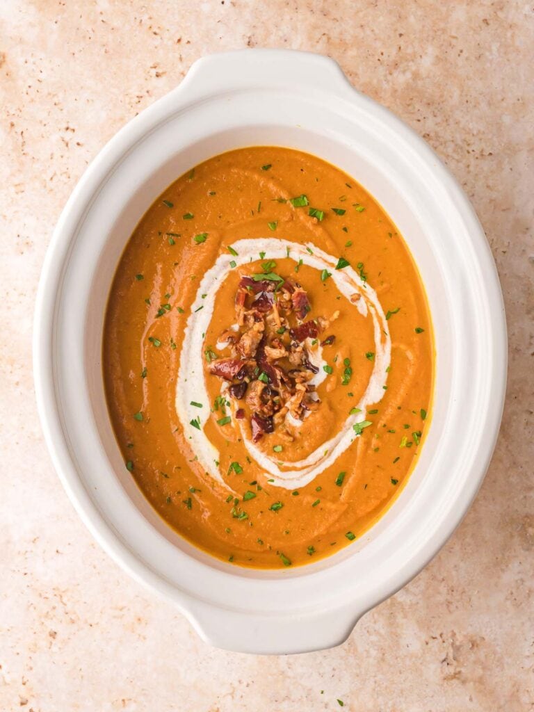 Overhead shot of creamy pumpkin soup in a white slow cooker, garnished with bacon and cream swirl.
