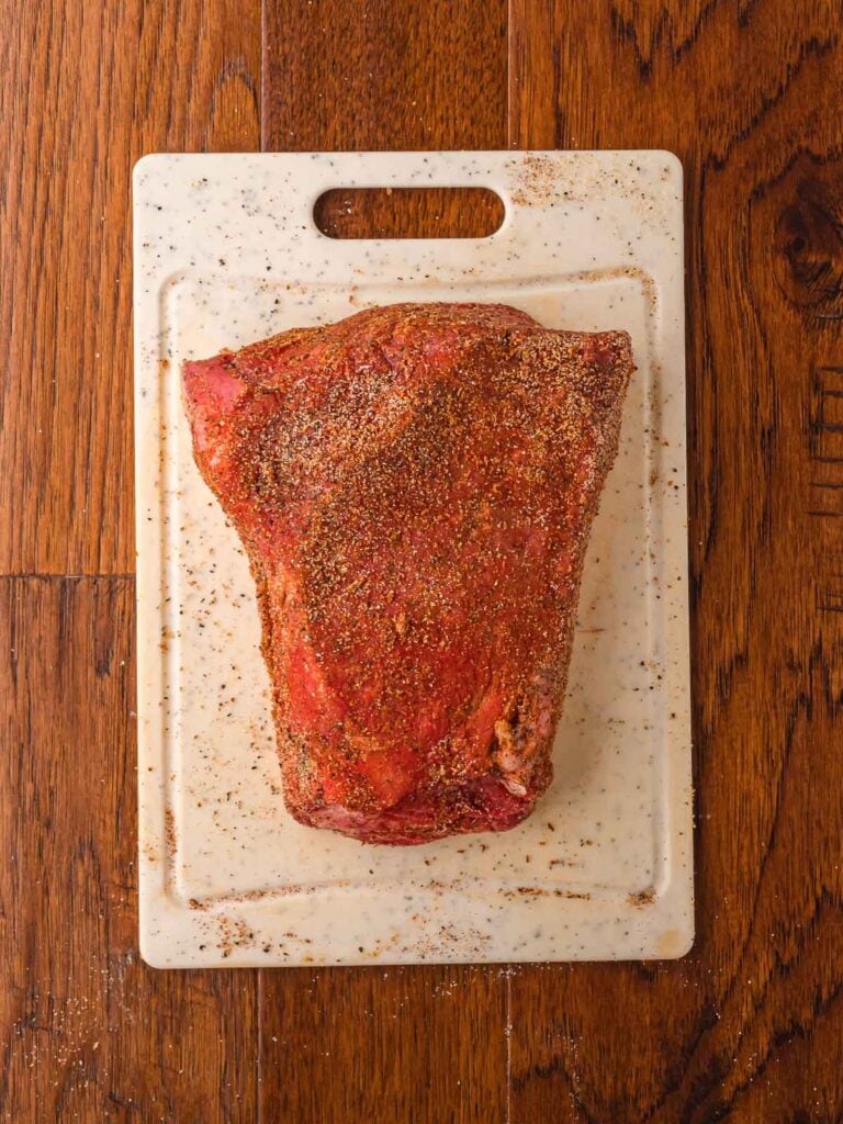 Raw chuck roast coated in a spice blend of cumin, paprika, chili powder, salt, and pepper on a cutting board.