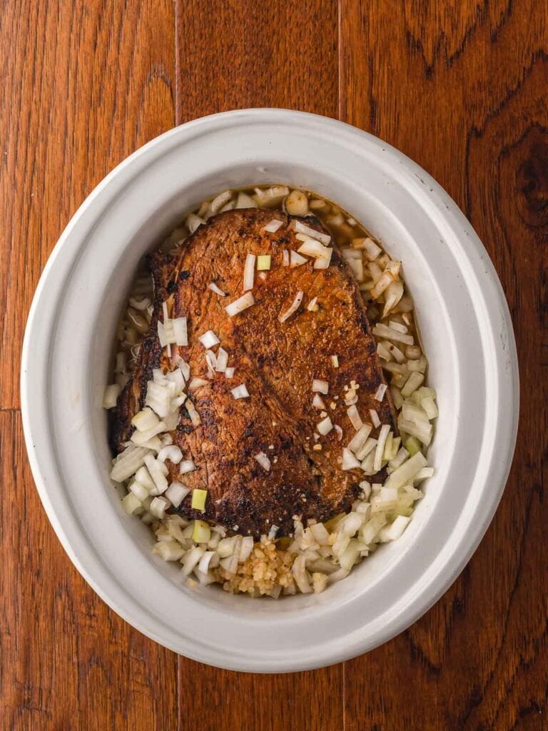 Seared chuck roast in a slow cooker surrounded by chopped onions and garlic.