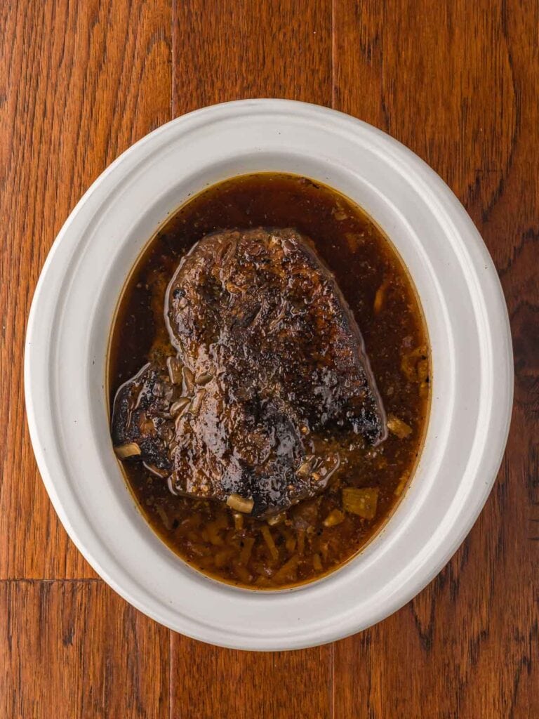 Chuck roast simmering in beef broth inside a slow cooker.