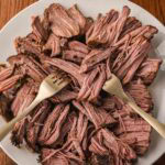 Tender slow cooked beef being shredded with two forks on a white plate.
