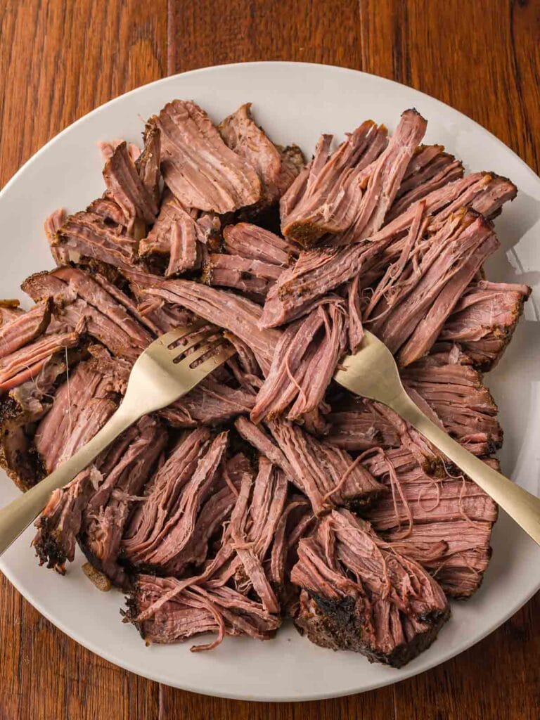 Tender slow cooked beef being shredded with two forks on a white plate.