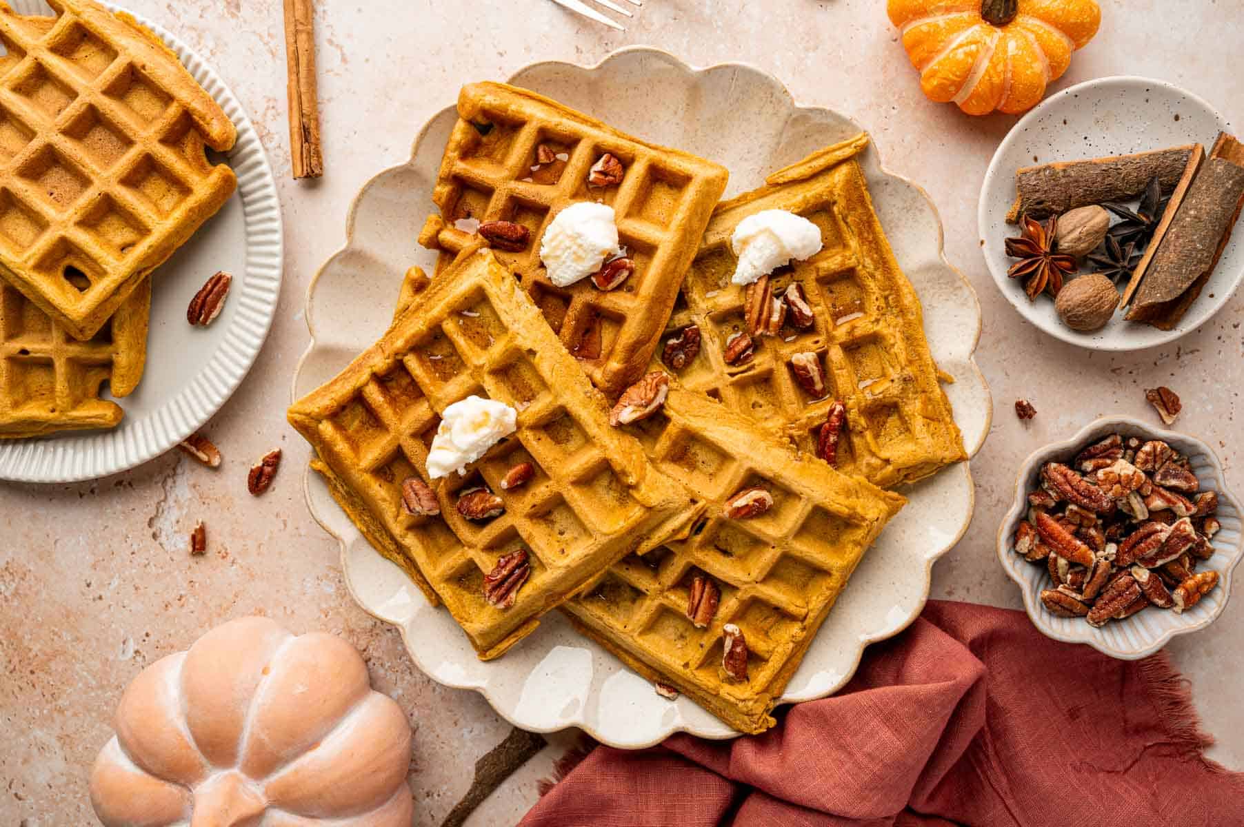 Golden pumpkin waffles topped with whipped cream and chopped pecans, served on a scalloped plate with warm spices and fall decor.