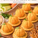 Mini pumpkin cheese balls on crackers with pretzel stems next to celery, carrots and shredded cheddar by Tessie's Table.