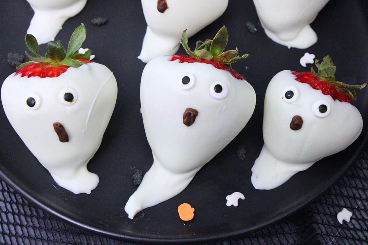 Fresh strawberries dipped in white chocolate and decorated with candy eyes and chocolate mouths to resemble little ghosts, placed on a black plate.