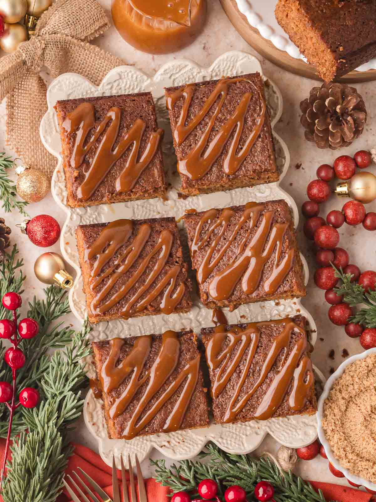 Festive display of Gingerbread Cake squares drizzled with caramel sauce on a serving platter surrounded by Christmas decorations.