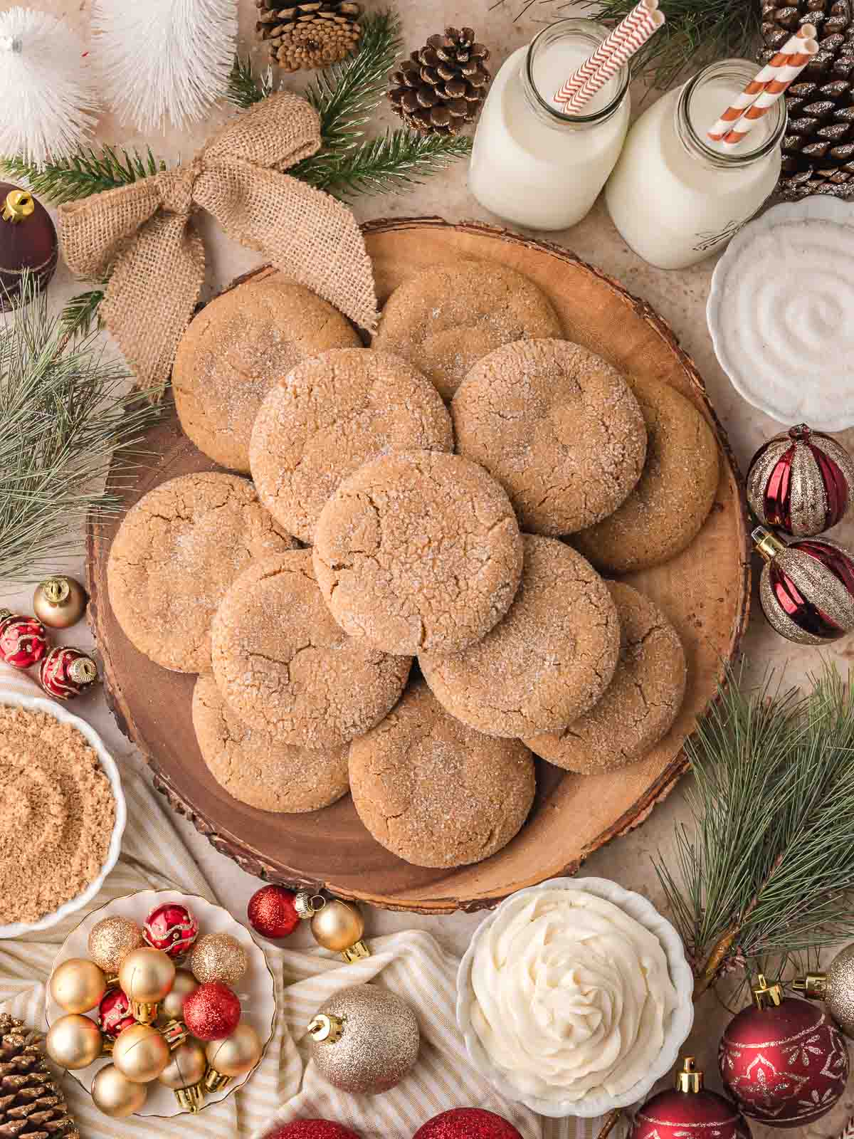 A festive plate of Gingerbread Cheesecake Cookies surrounded by holiday decorations, pine branches, and milk jars with striped straws.
