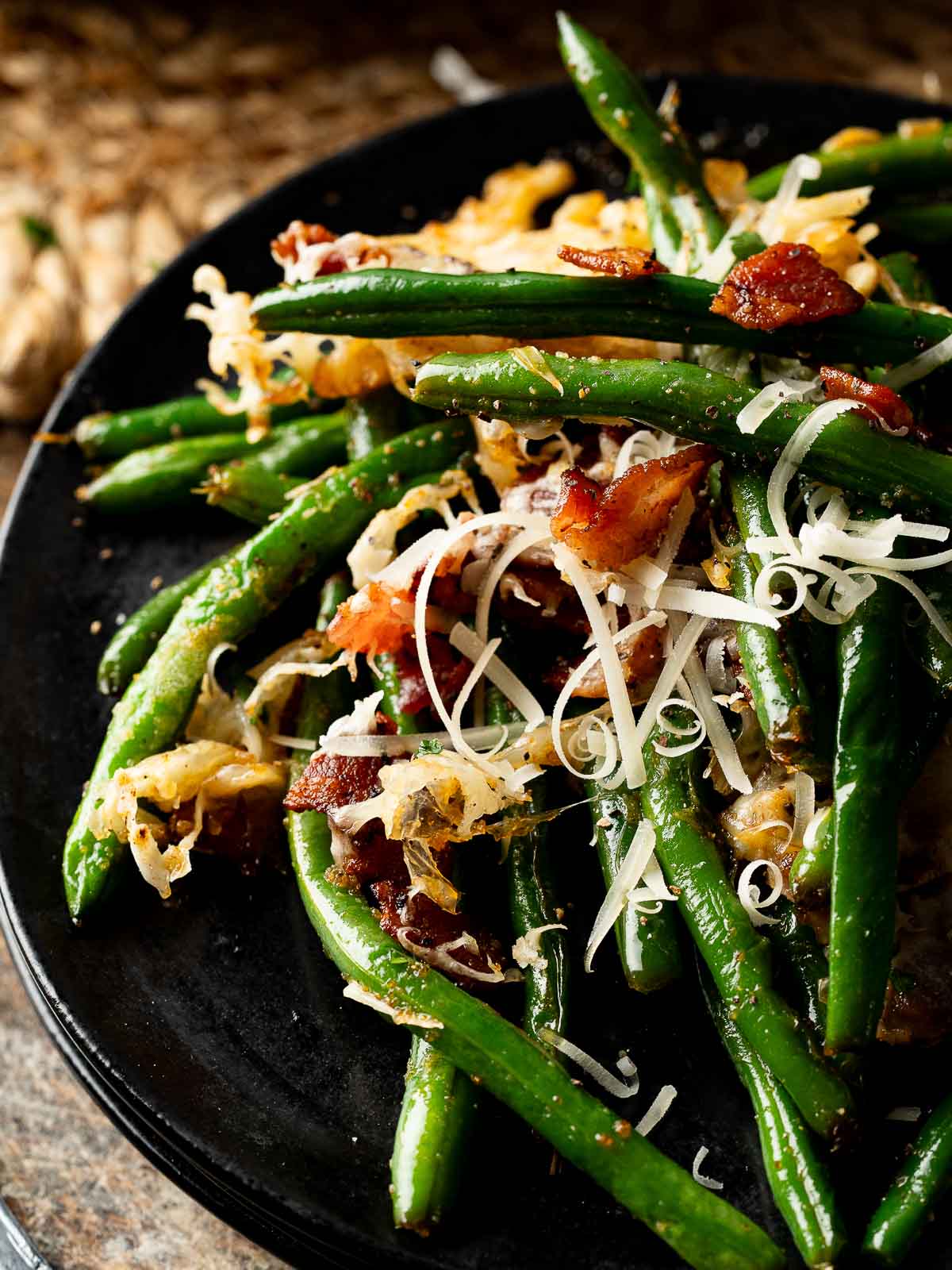 Close-up of cheesy green beans with bacon and Parmesan served on a black plate.