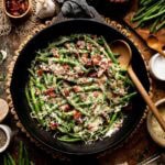 Cheesy green beans in cast iron skillet surrounded by fresh ingredients on table.