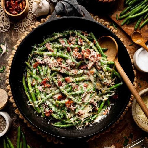 Cheesy green beans in cast iron skillet surrounded by fresh ingredients on table.