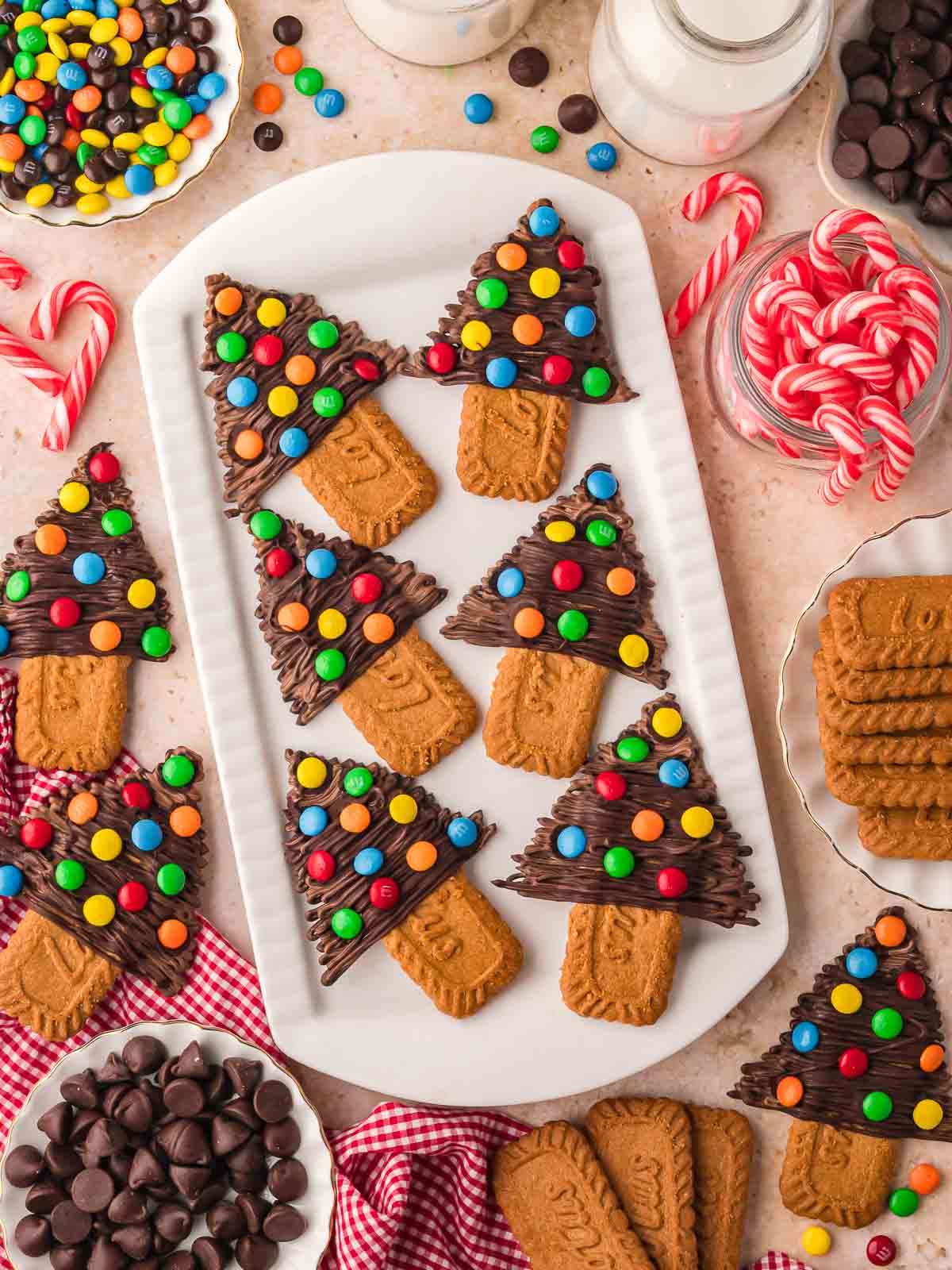 Chocolate Christmas Trees arranged on a white platter with Biscoff cookies, candy canes, and bowls of chocolate chips and colorful candies on the table.