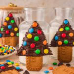 Close-up of Chocolate Christmas Trees standing against jars of milk with colorful M&M decorations and scattered candies on the table.