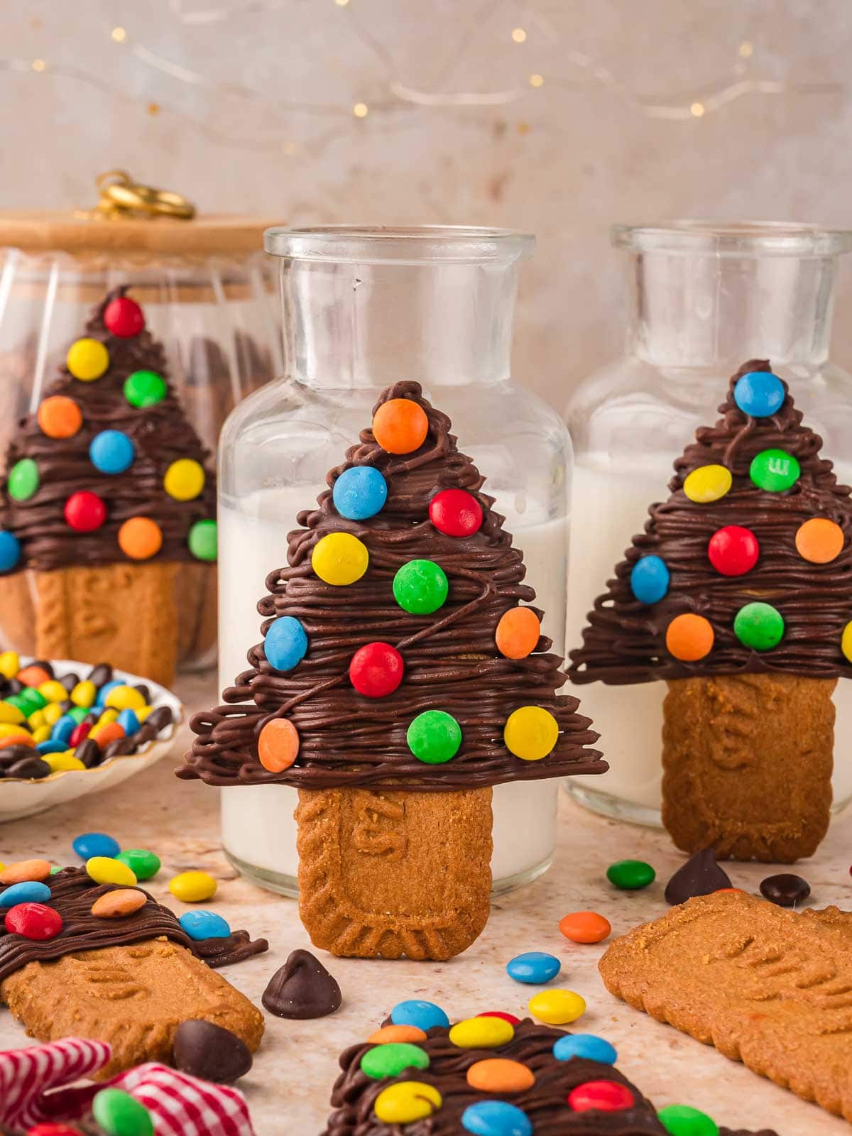 Close-up of Chocolate Christmas Trees standing against jars of milk with colorful M&M decorations and scattered candies on the table.