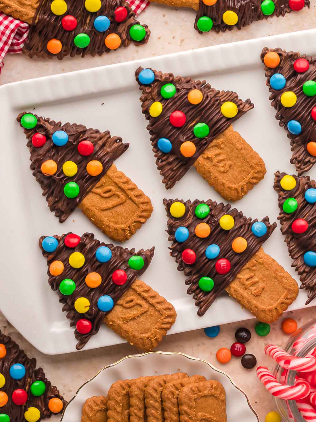 Top view of Chocolate Christmas Trees made with Biscoff cookies, melted chocolate, and mini M&Ms displayed on a white serving tray with festive red checkered napkin and candy canes.