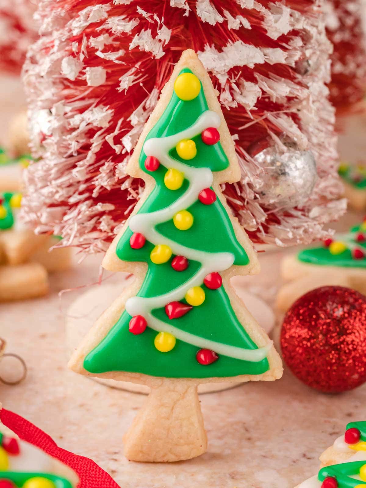 Decorated Christmas tree sugar cookie with green icing and colorful ornaments in front of red holiday decorations.