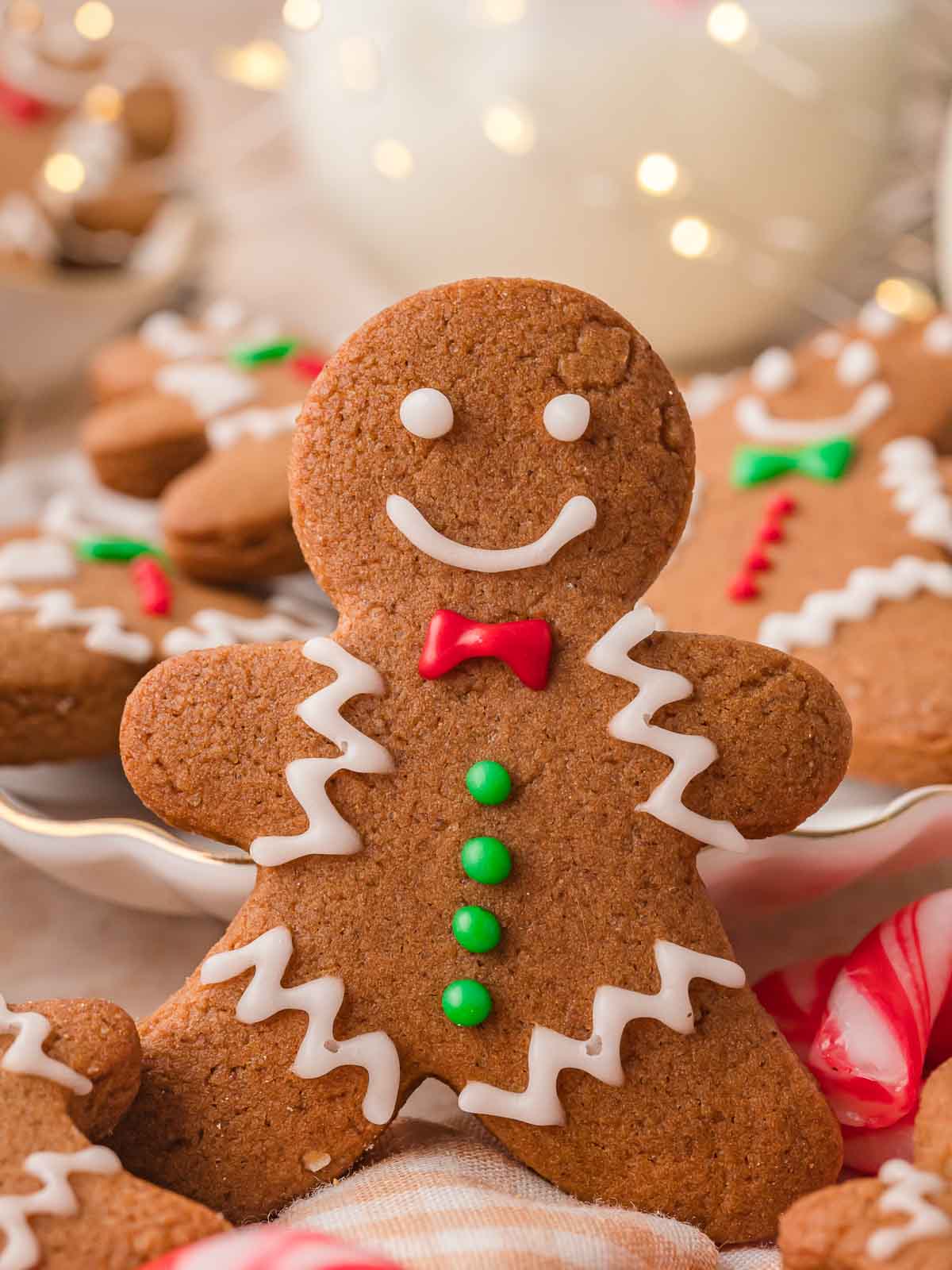 Close-up of a smiling Gingerbread Man Cookie with red bow tie and green buttons, surrounded by festive holiday treats.