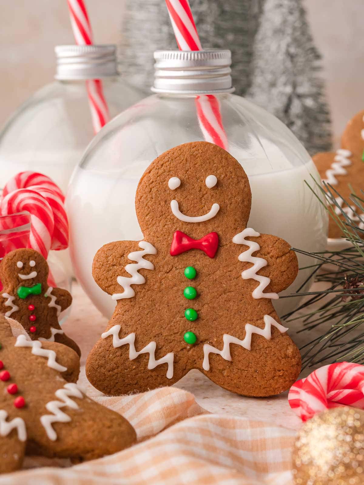 Gingerbread Man Cookie standing in front of bottles of milk with red and white straws, candy canes, and Christmas greenery in the background.