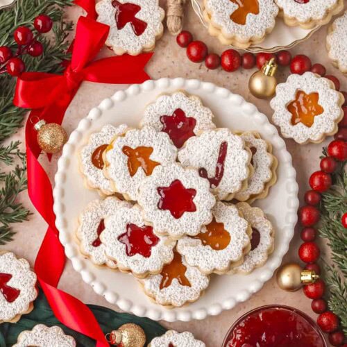 Jam-filled Linzer cookies arranged with red bows, cranberries, and gold ornaments for a Christmas spread.