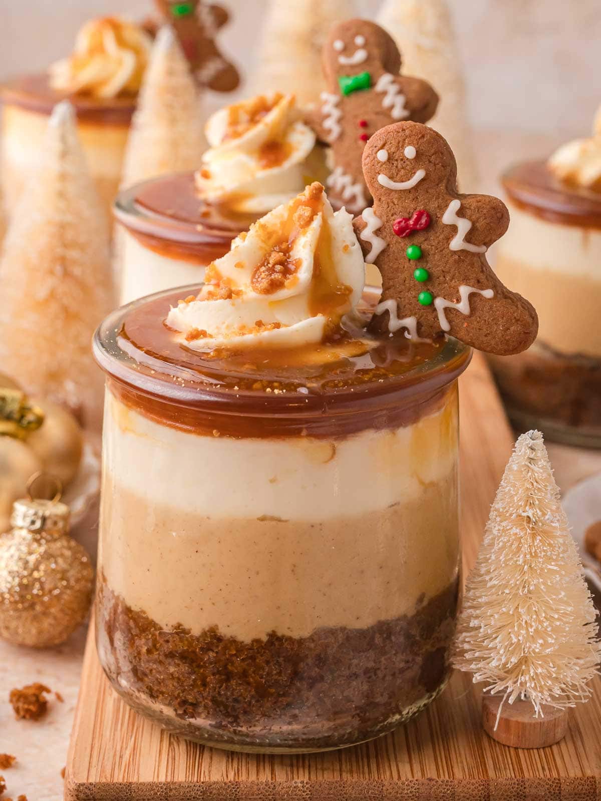 Holiday display of Mini Gingerbread Trifles surrounded by small Christmas trees and ornaments, showing layered dessert jars with gingerbread men on top.
