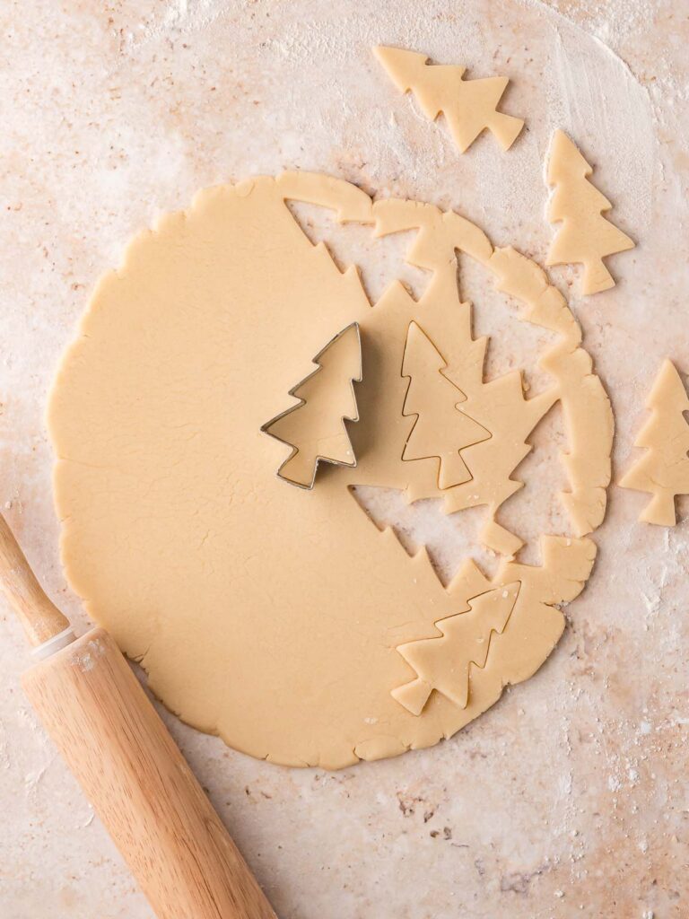 Rolled-out cookie dough with Christmas tree shapes being cut out using a metal cookie cutter.