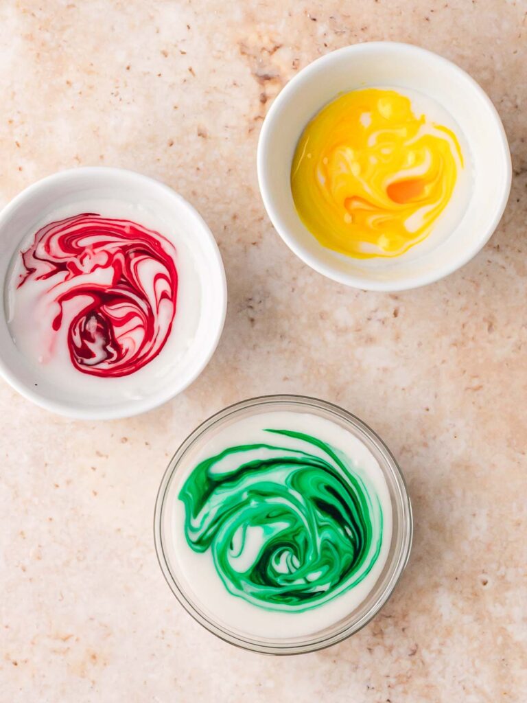Small bowls of glaze icing tinted red, yellow, and green for decorating Christmas sugar cookies.