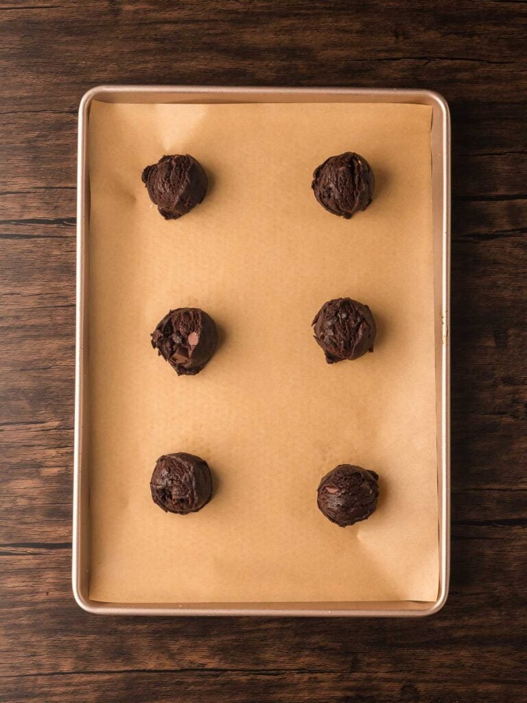 Scoops of Double Chocolate Chunk Cookie dough spaced on a parchment-lined baking sheet before baking.