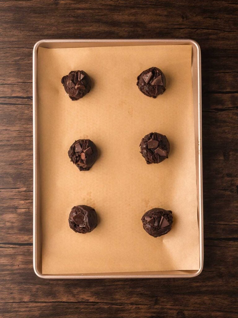 Cookie dough balls topped with extra dark chocolate chunks on a parchment-lined baking tray ready for the oven.