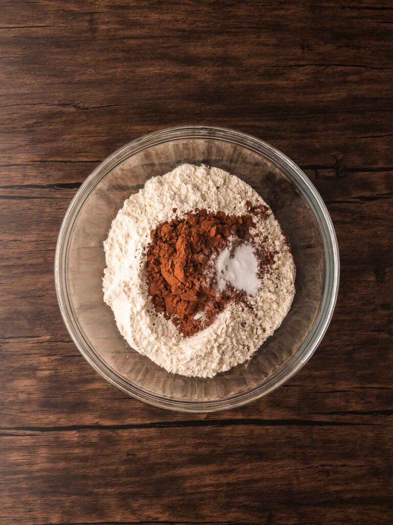 Dry ingredients for Double Chocolate Chunk Cookies in a glass bowl with flour, cocoa powder, baking soda, and salt on a wooden surface.