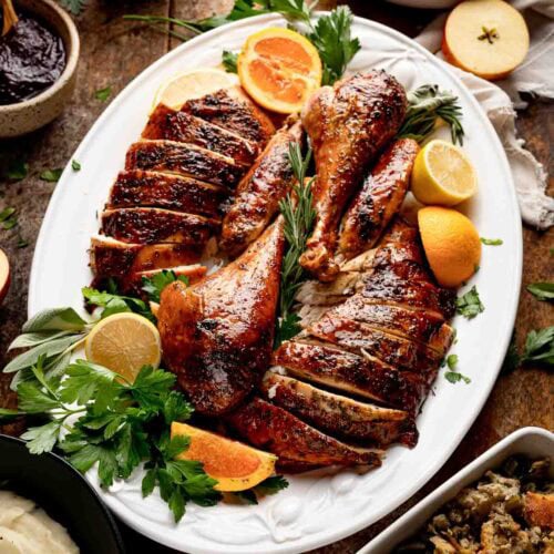 Sliced roast turkey arranged on white platter with oranges, lemons, and herbs surrounded by holiday side dishes.