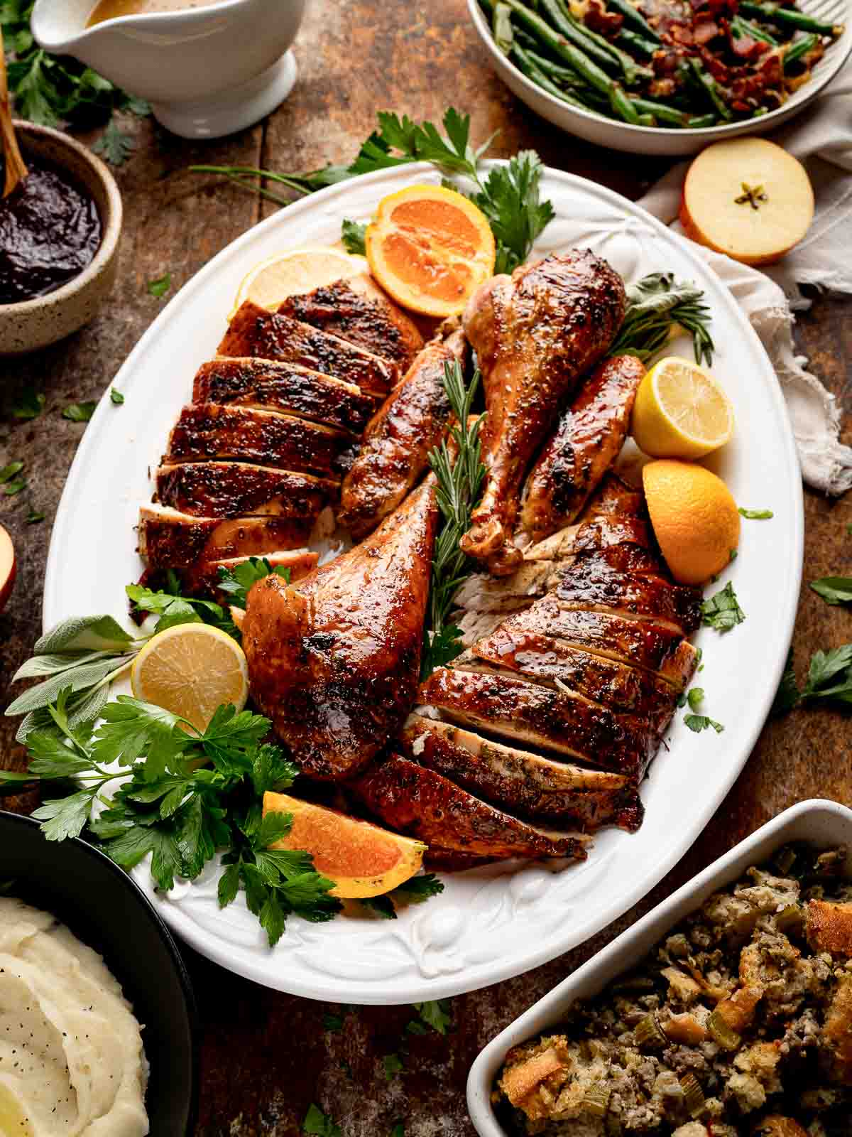 Sliced roast turkey arranged on white platter with oranges, lemons, and herbs surrounded by holiday side dishes.