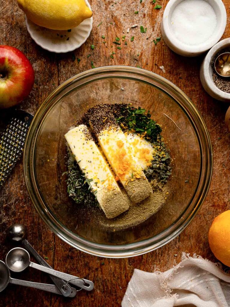 Softened butter mixed with chopped herbs, lemon zest, and seasonings in a glass bowl for herb butter roast turkey.