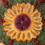 Golden puff pastry sausage roll wreath shaped like a Christmas wreath with a bowl of cranberry sauce in the center, surrounded by greenery, red berries, and festive ribbon decorations.