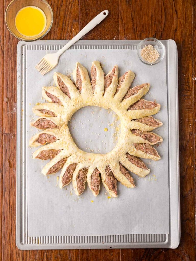 Unbaked sausage roll wreath brushed with egg wash and sprinkled with sesame seeds.
